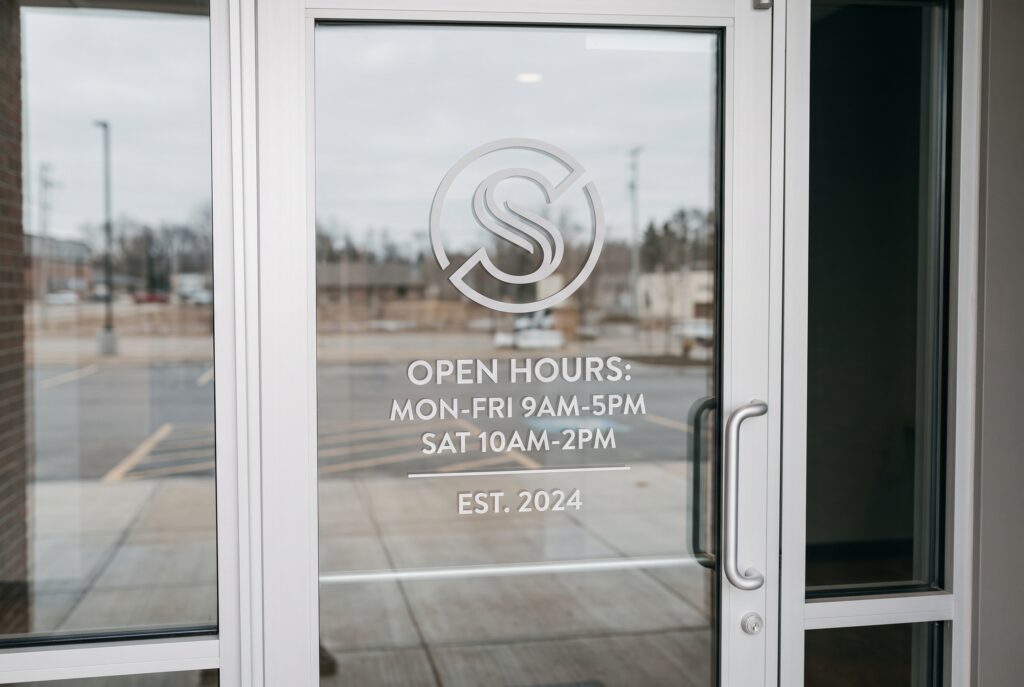 A close-up view of a clear glass front entry door on a modern commercial building. The door features a custom etched "S" logo and detailed store hours: "OPEN HOURS: MON-FRI 9AM-5PM, SAT 10AM-2PM. EST. 2024," providing a professional first impression.