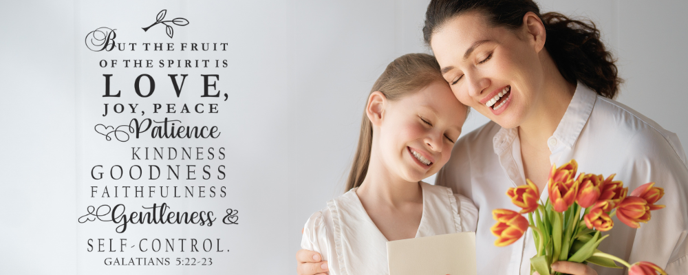A mother and daughter hugging after a gift of a scripture vinyl wall decal from The Simple Stencil - the bible verse decal is from the verse Galatians 5:22 and is shown on a white wall. 