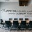 A sleek conference room wall featuring a large, inspiring wall decal: "“TEAMWORK is the ability to work together toward a COMMON VISION.” -Andrew Carnegie". A long wooden meeting table with black ergonomic chairs is set in the foreground, with natural light coming from a vertical window.